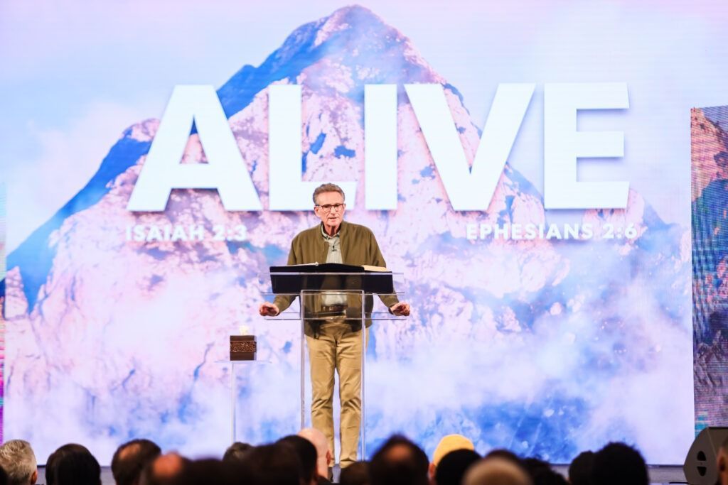 Speaker at podium on AIVE conference stage with mountain backdrop and audience in foreground