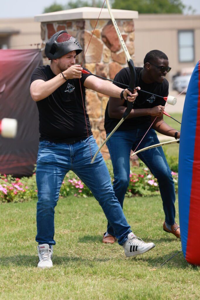 Two people aiming bows outdoors during an archery game, wearing black shirts and jeans.