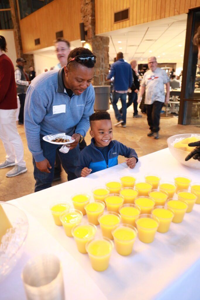 Child and adult sampling yellow drinks at a table in a hall