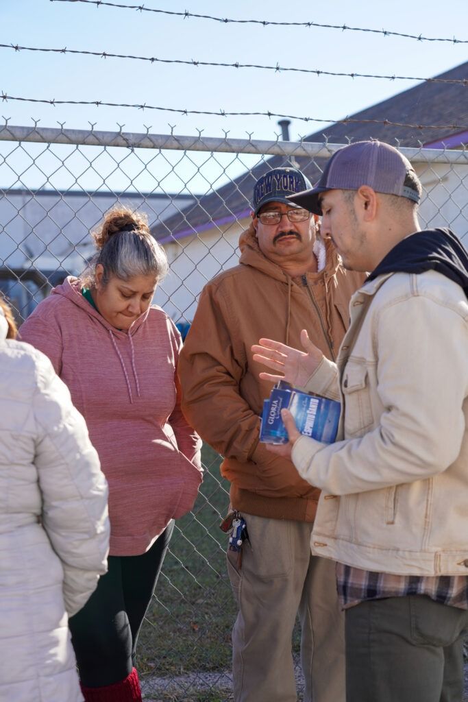 People standing outdoors by a chain-link fence, talking near a house in daylight.