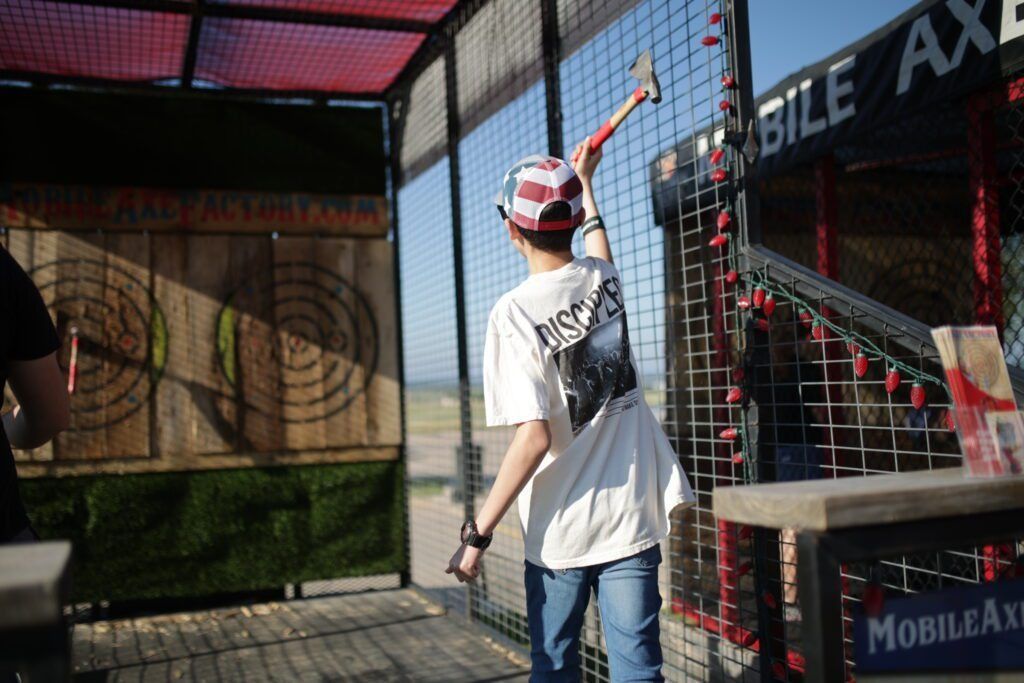 Person in a red cap aims a paintball marker at an outdoor target range.