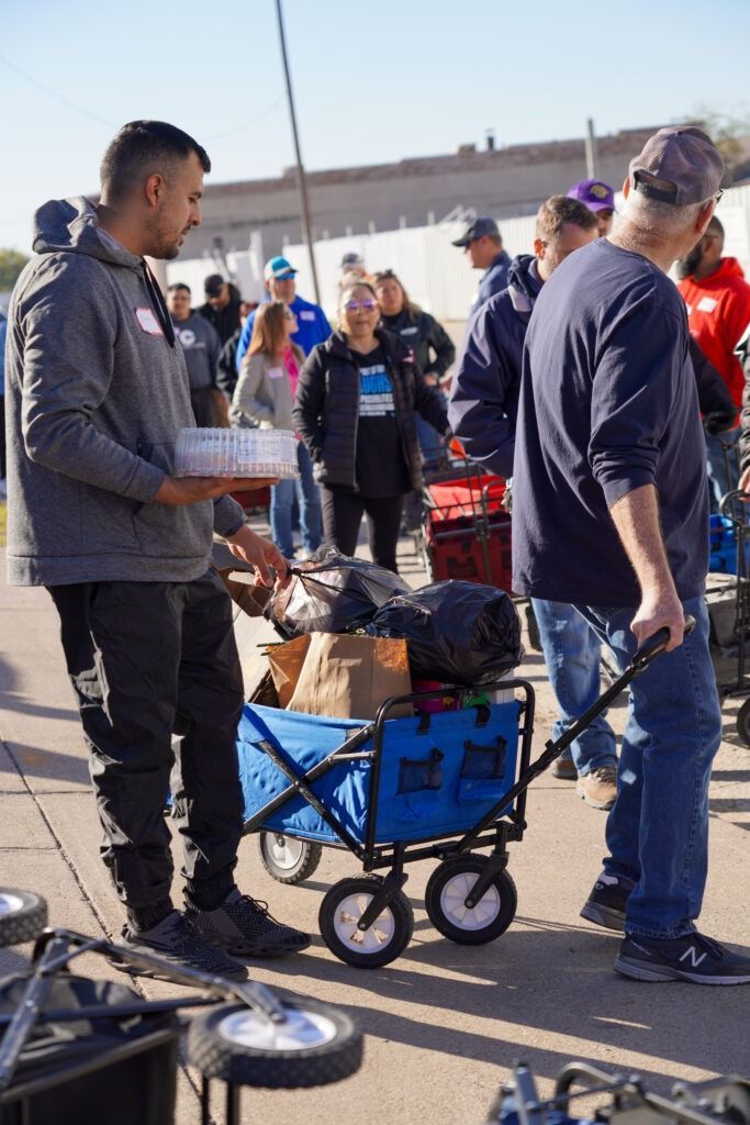 People gathered outdoors around a blue wagon loaded with bags and boxes at an event