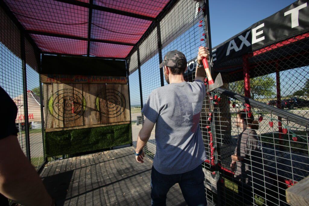 Person standing at an axe-throwing lane, raising an axe toward the target behind a mesh cage.