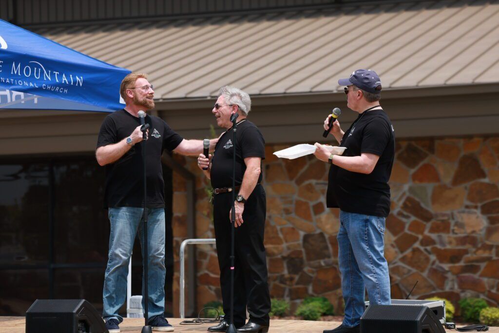 Three people in black shirts stand on an outdoor stage with microphones, speaking to an audience.