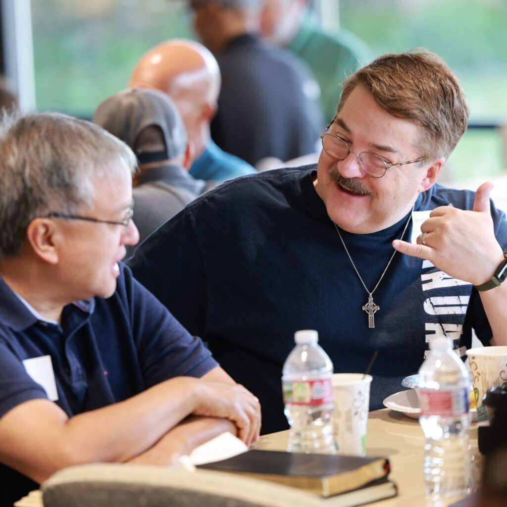 Two men chatting at a table with drinks and a book, one smiling and wearing a cross necklace