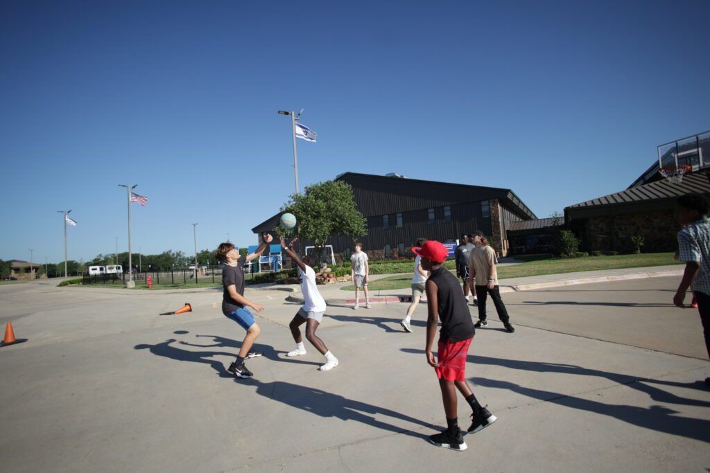 People skating on a sunny open plaza near low buildings and trees.