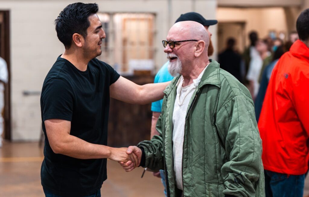 Two men shaking hands indoors, one in a black shirt and one in a green jacket, smiling.