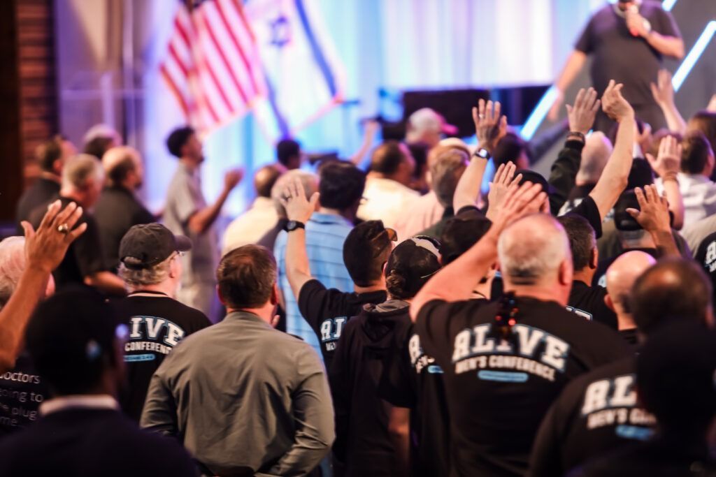 Crowd with raised hands at an indoor event, facing a stage with an American flag in the background