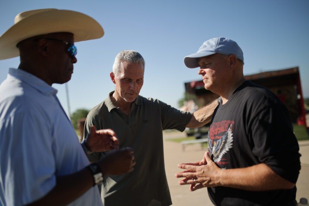 Three men talking outdoors in bright sunlight, one in a cowboy hat and one in a cap.