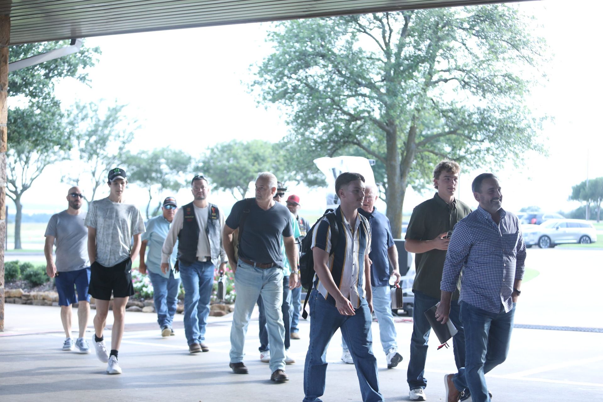 A group of people walking under a covered outdoor walkway near trees and parked cars.