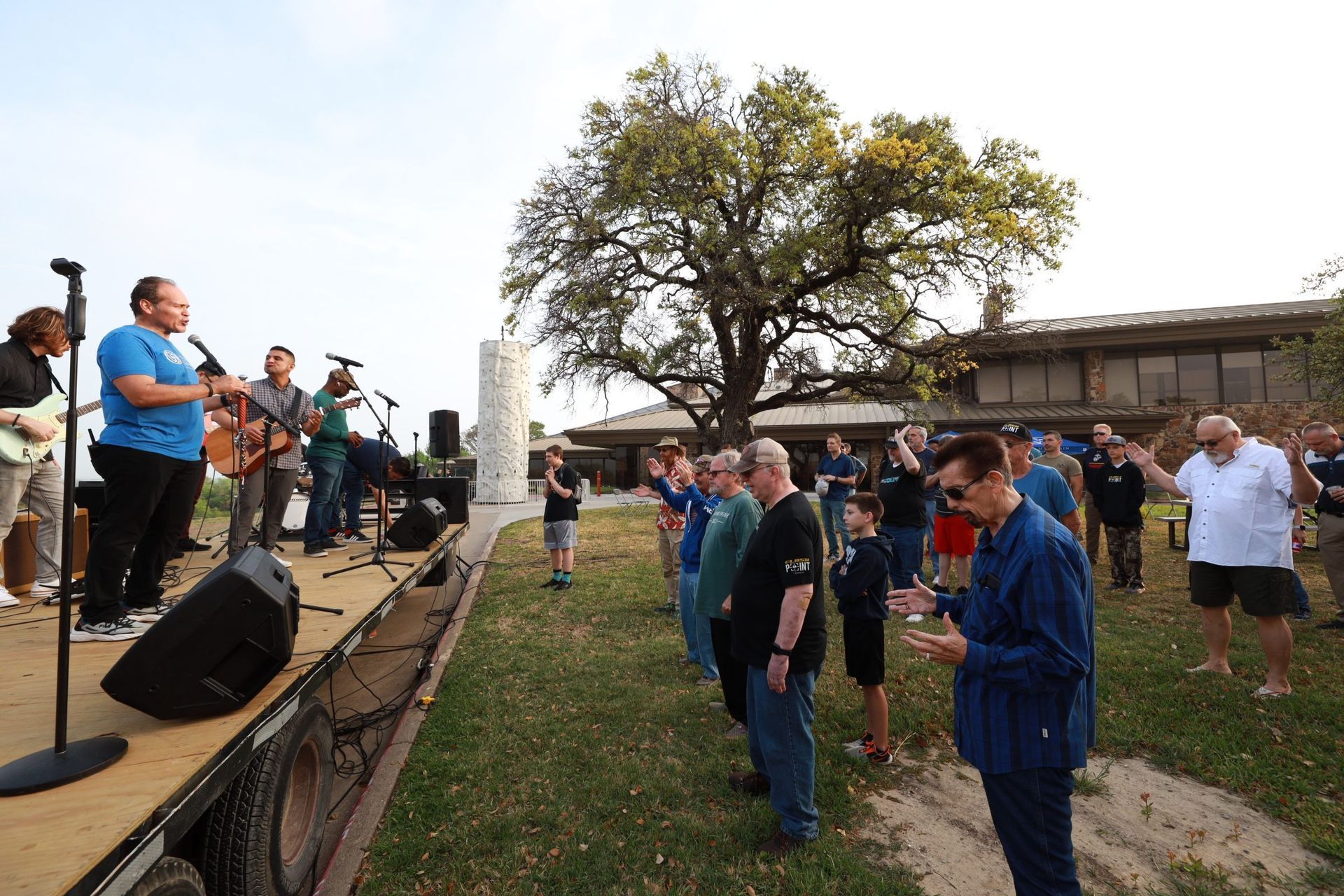 Musicians perform on a small outdoor stage while an audience stands on the grass nearby.