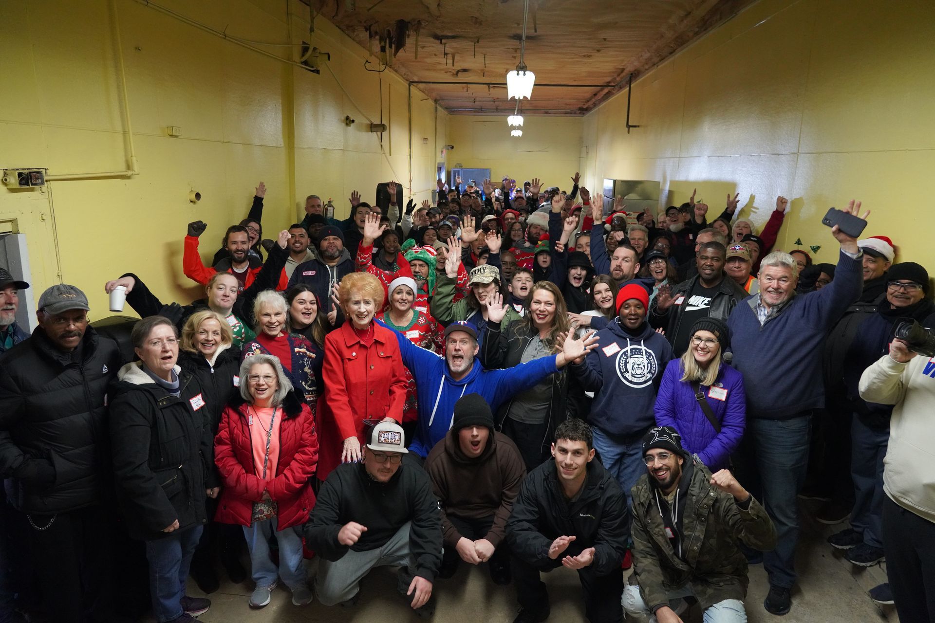 Large group posing indoors in a hall, smiling and waving, with red and dark winter jackets.