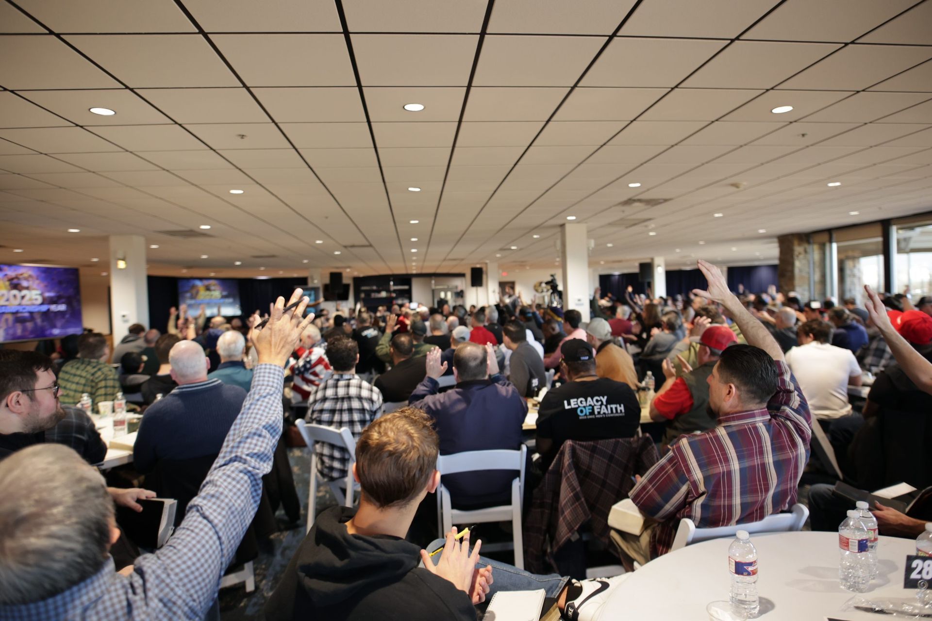 Crowded conference room with people seated at tables, many raising hands during a presentation