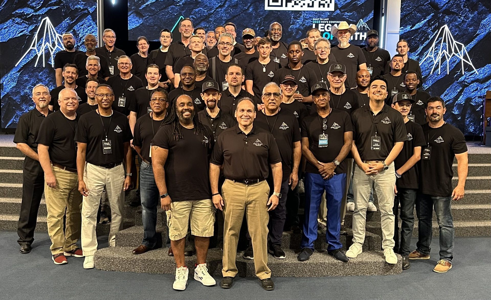 Large group posing for a team photo indoors in front of a stage backdrop, wearing matching black shirts.
