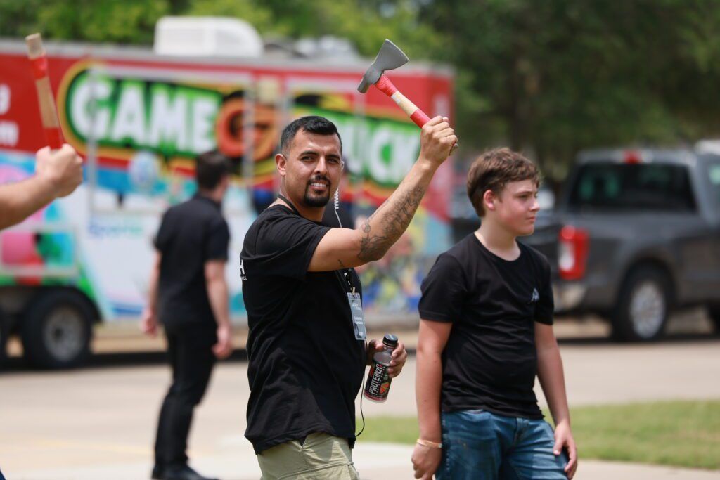 Two men in black shirts walking outdoors, one raising an axe-shaped object, with a colorful truck in the background