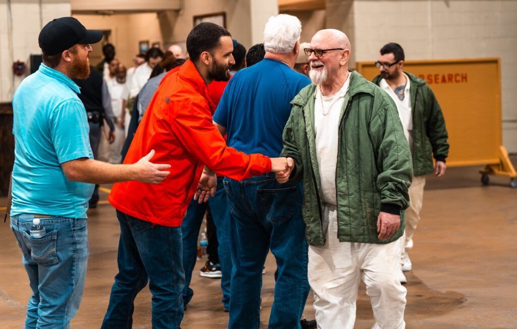 Two men in red and teal greet an older man in a green jacket at an indoor event.