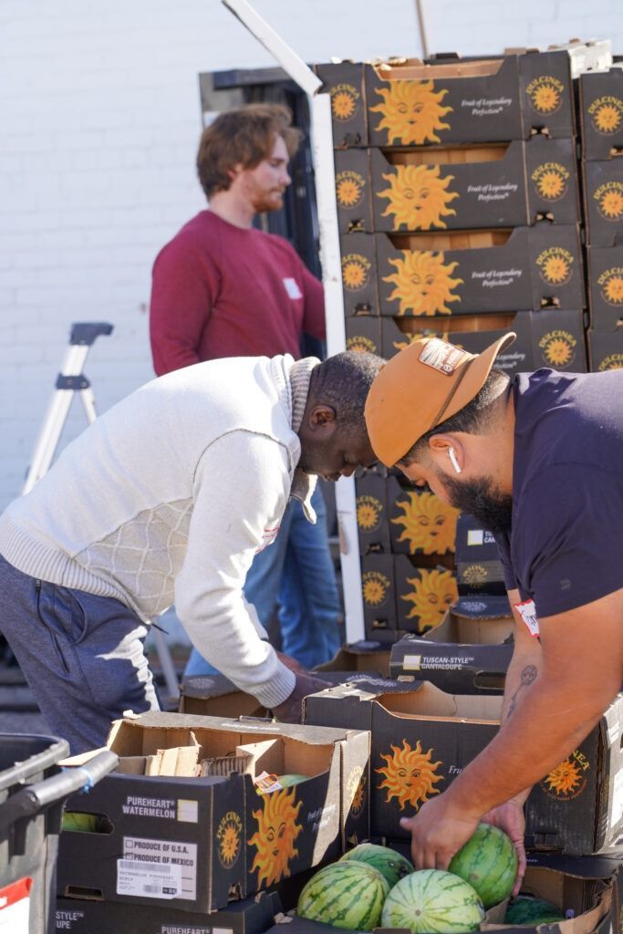 Volunteers packing boxes of produce, including watermelons, beside stacked crates outdoors