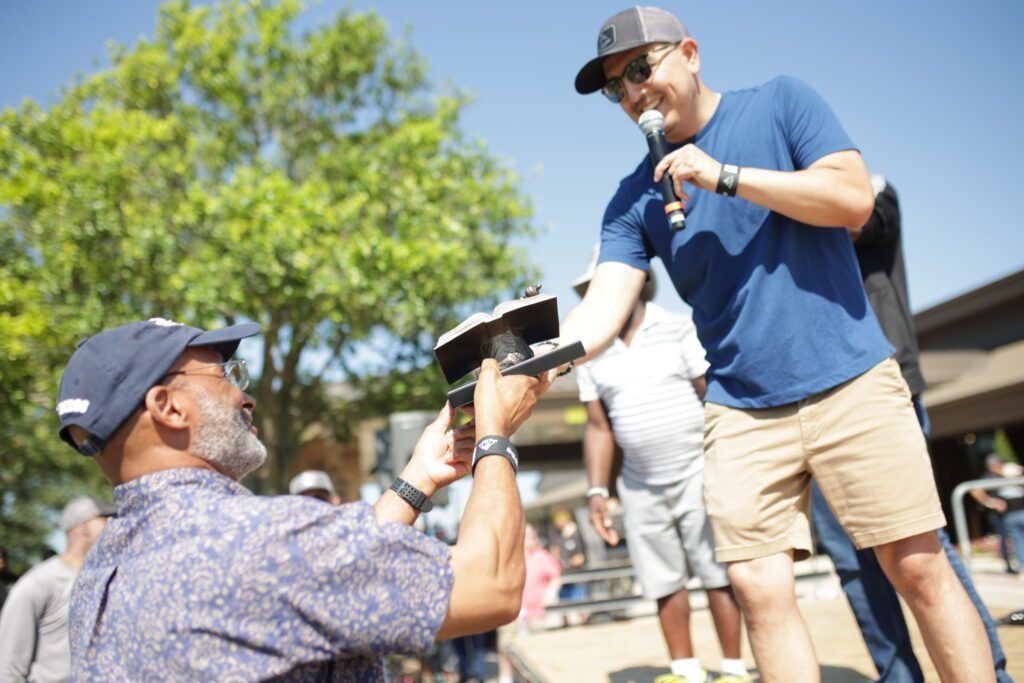 Two men outdoors, one kneeling and holding a device while the other smiles and gestures beside him.