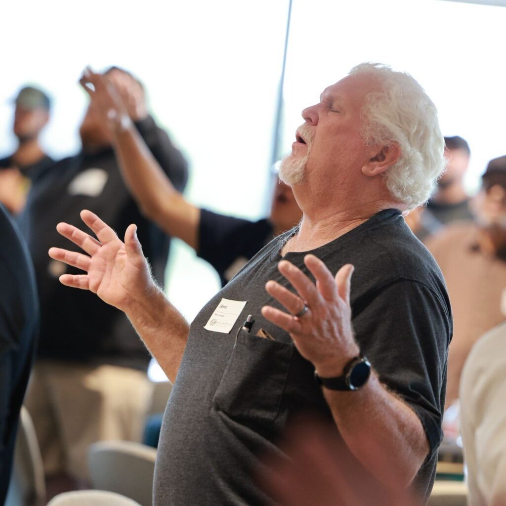 Man speaking passionately with raised hands at a meeting, others blurred in the background.