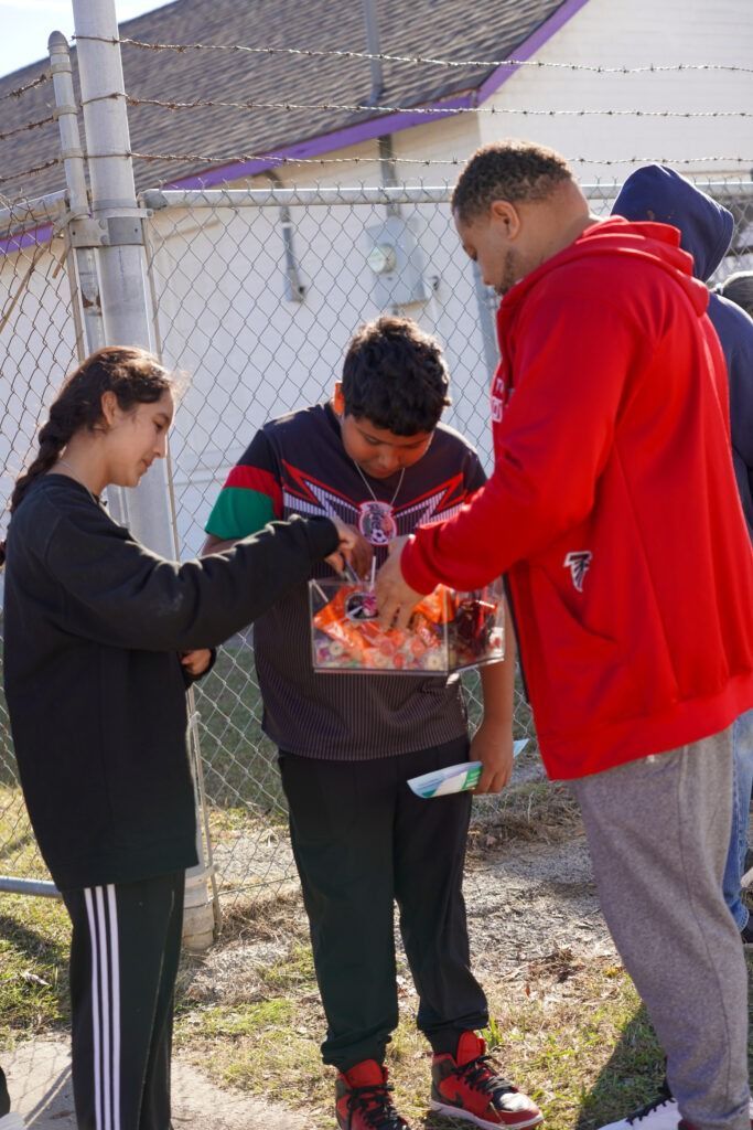 Three people stand outdoors near a fence, looking at a box of snacks or toys.