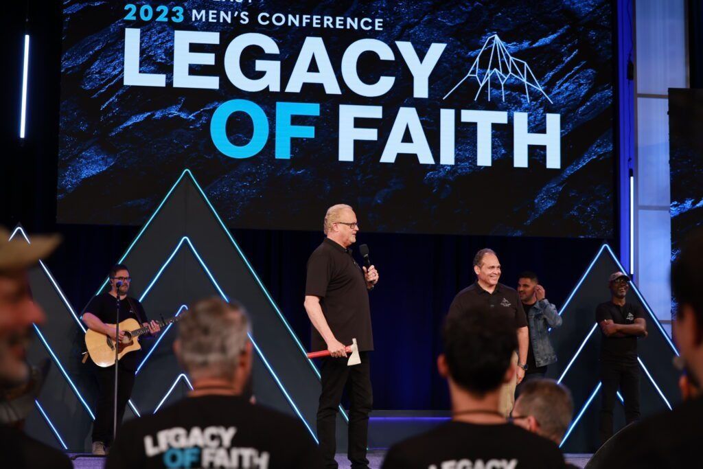 Conference stage with “Legacy of Faith” backdrop, speakers under blue lights, audience in foreground