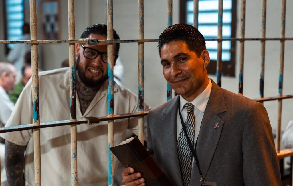 Two men behind metal bars, one in a gray suit holding a folder, in an indoor hallway