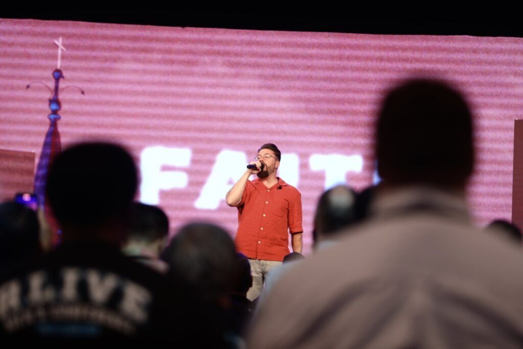 Speaker on stage addressing a crowd, with blurred audience silhouettes in the foreground