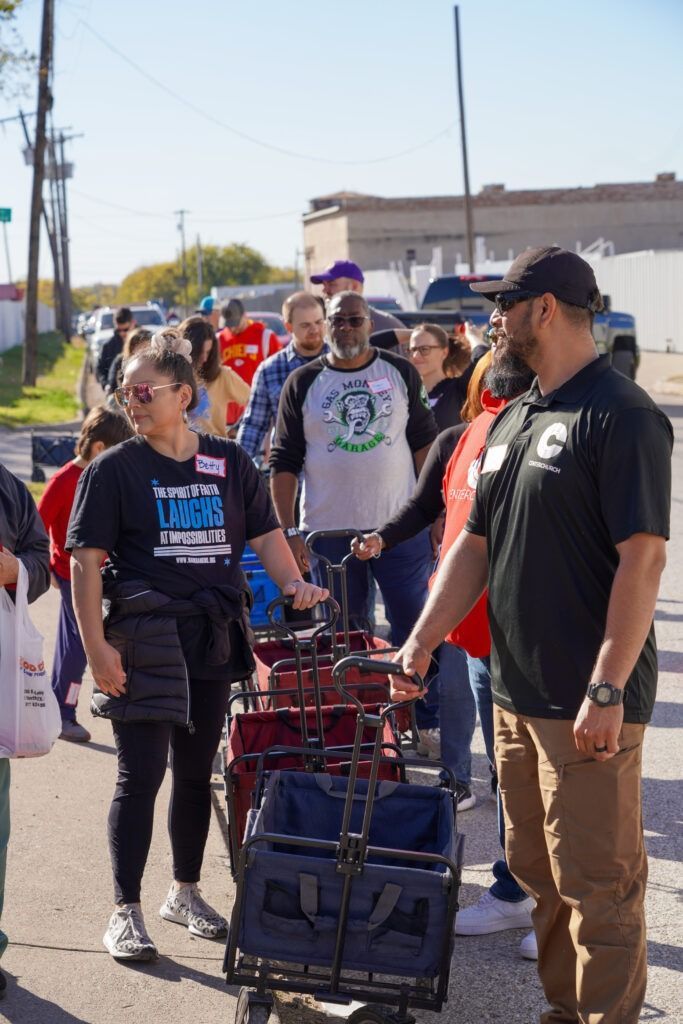 People standing in line outdoors, with a man in a black jacket beside stacked blue crates.