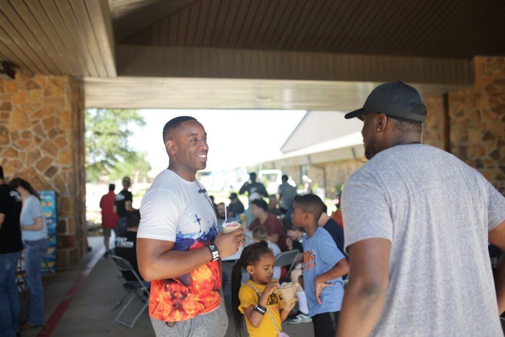 People standing and talking in a covered outdoor area, with children in the background.