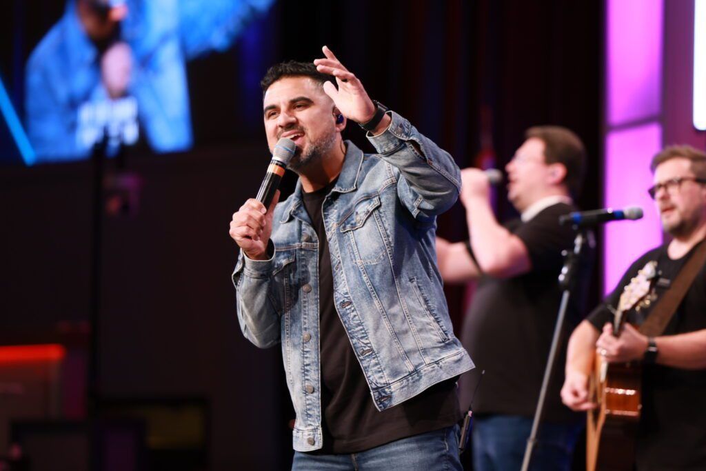 Singer in denim jacket performing on stage with microphone, band playing in background