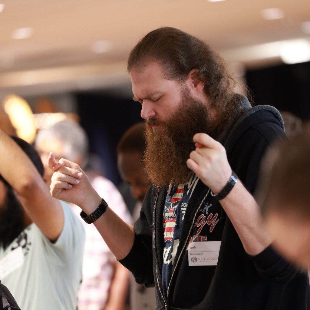 Bearded man with eyes closed, hands raised in a crowd at an indoor event