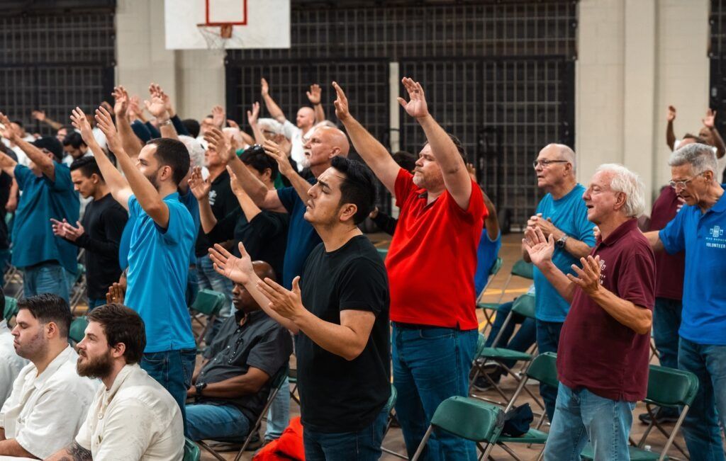 Audience standing and clapping indoors, with arms raised in a gym-like venue.