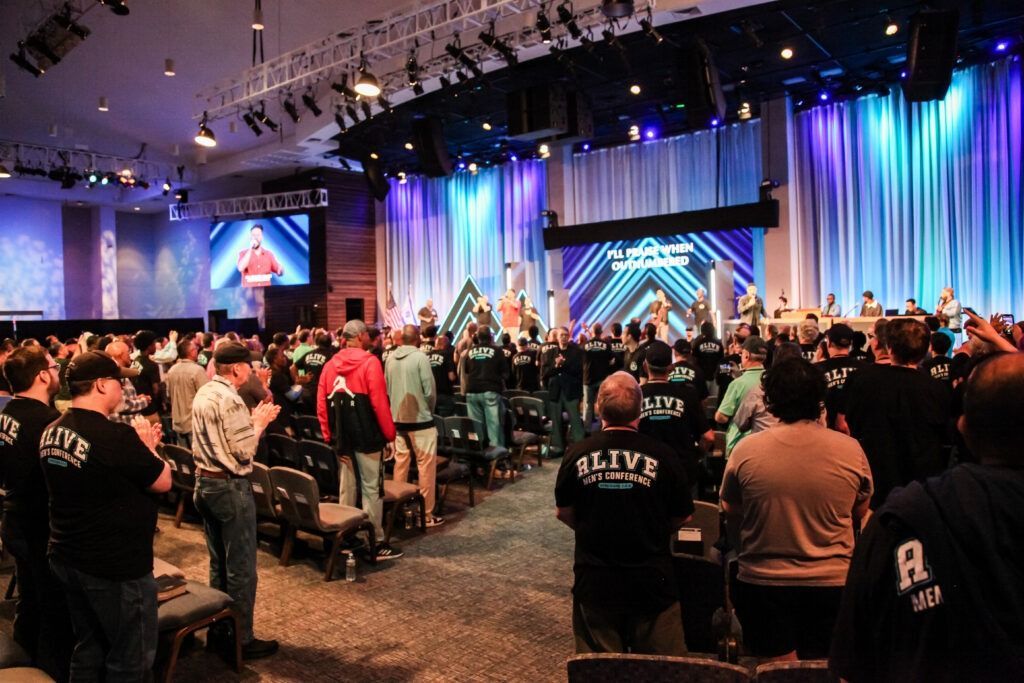 Conference audience facing lit stage with blue and purple lights and large screens