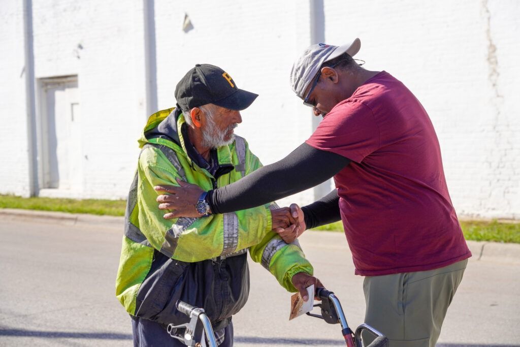 Two men outdoors shaking hands, one in a bright safety vest and the other in a maroon shirt.