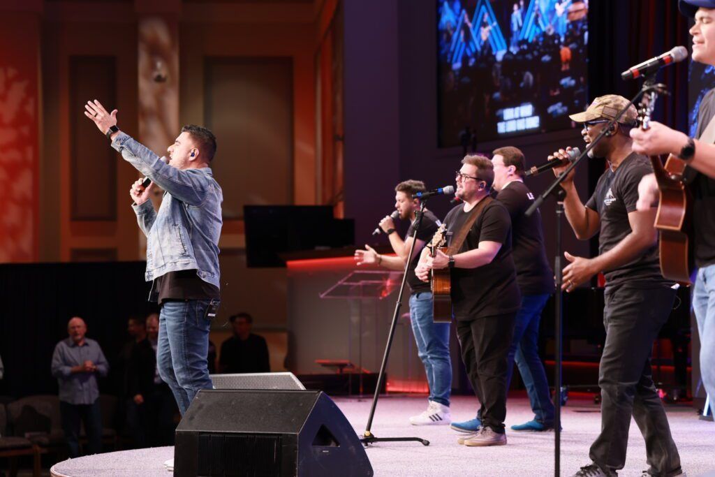 Musicians performing on stage in a church, with one singer raising his arms and others playing instruments.