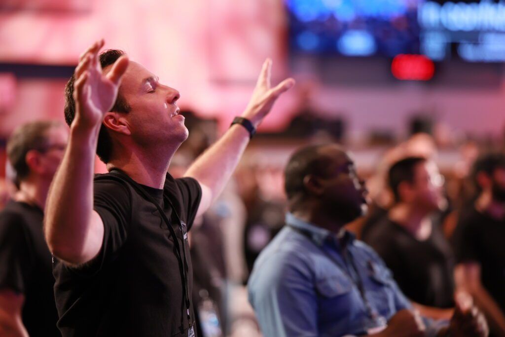 Two people in a crowd, one looking up with hands raised in a brightly lit indoor venue