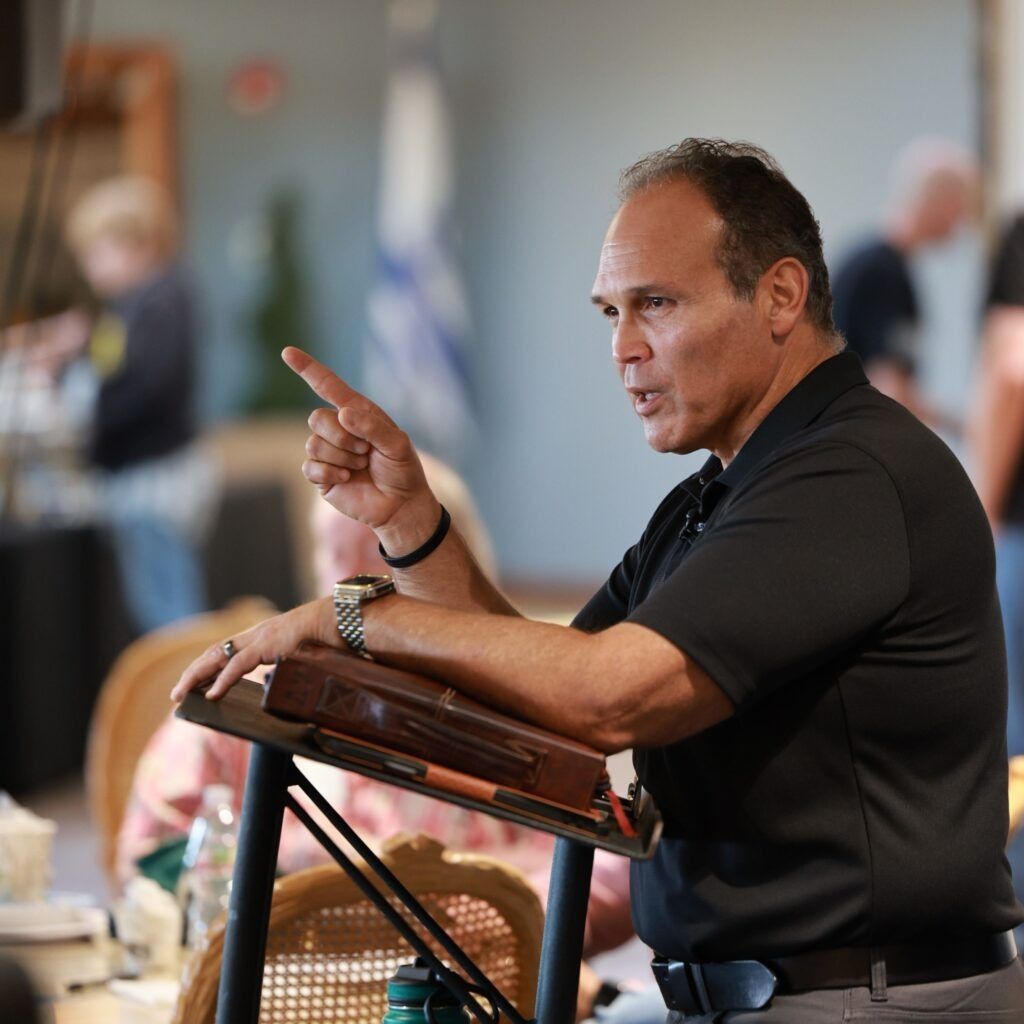Man speaking at a podium, pointing emphatically during a presentation in a blurred indoor setting