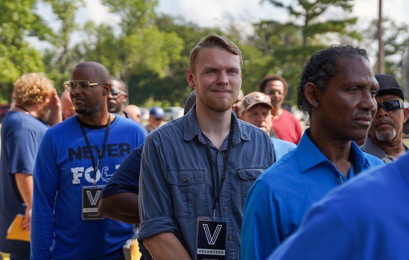 People standing outdoors in a line, including a man in a blue shirt and a smiling man in a denim shirt with a lanyard