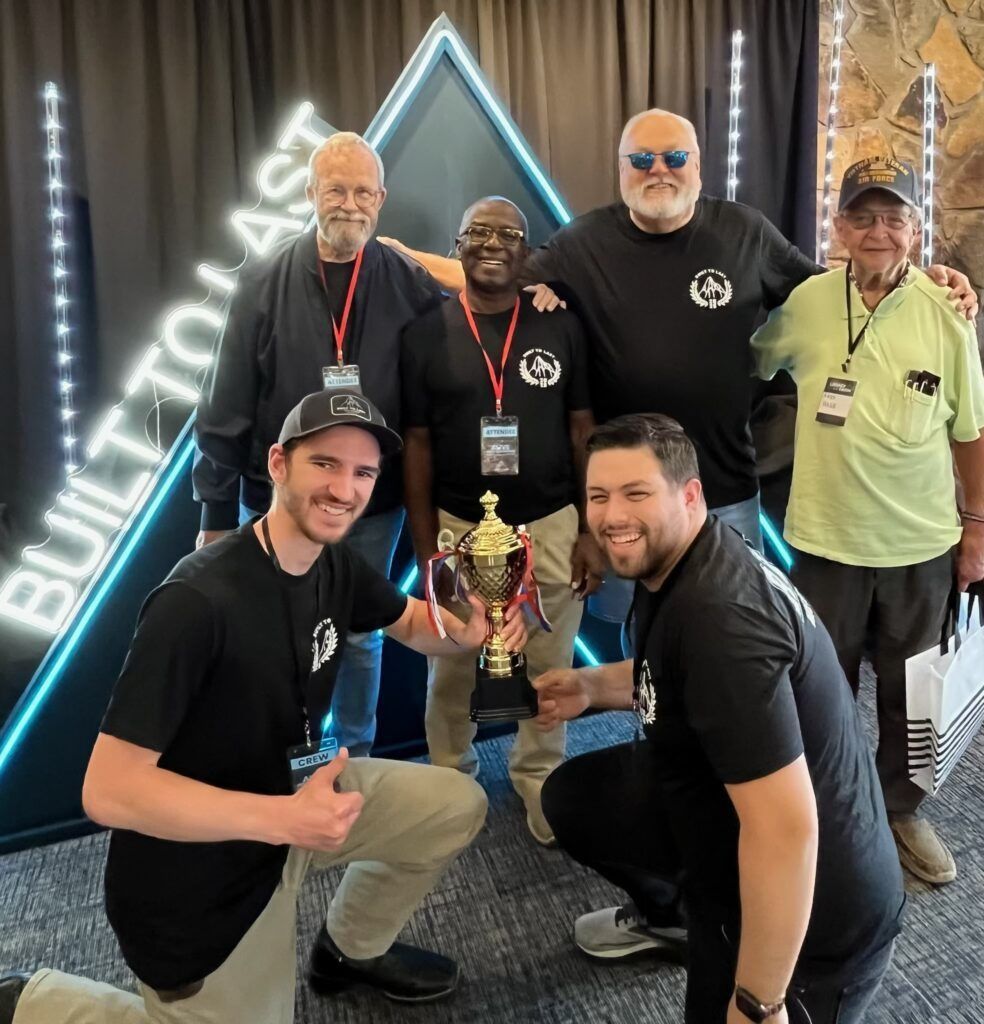 Group of five smiling people posing with a trophy at an indoor event booth.