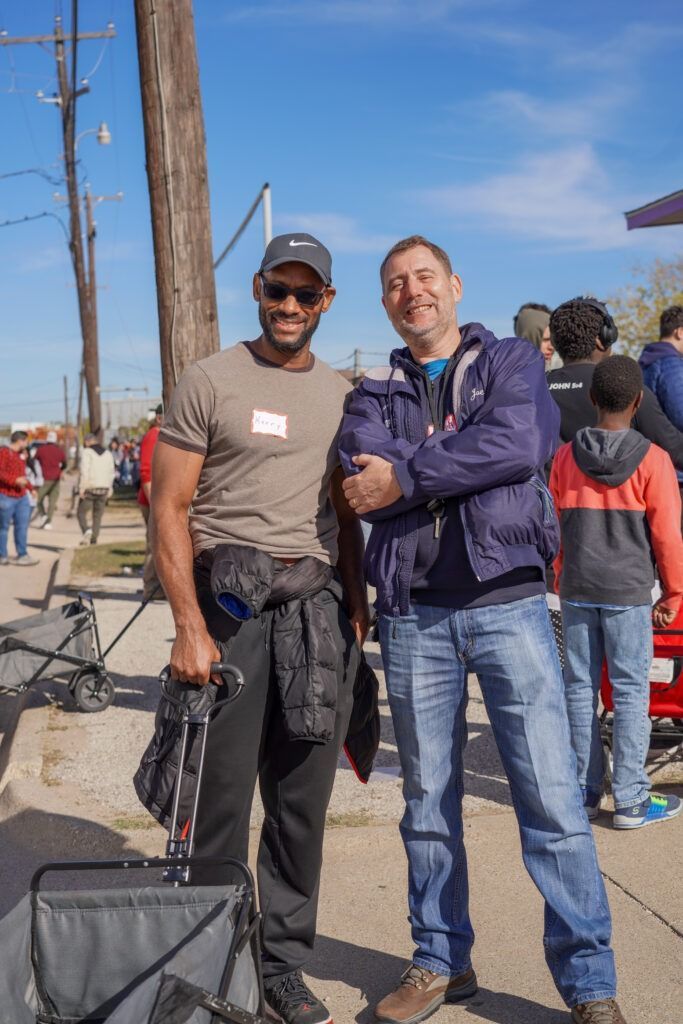 Two men posing together on a sunny street, one holding luggage and wearing a cap and sunglasses.