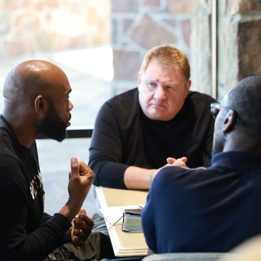 Three people in a meeting around a table, talking with laptops and papers visible.