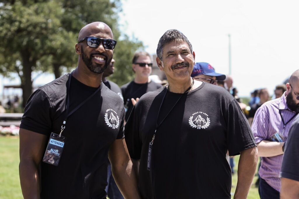 Two men in black T-shirts smiling outdoors at a sunny event