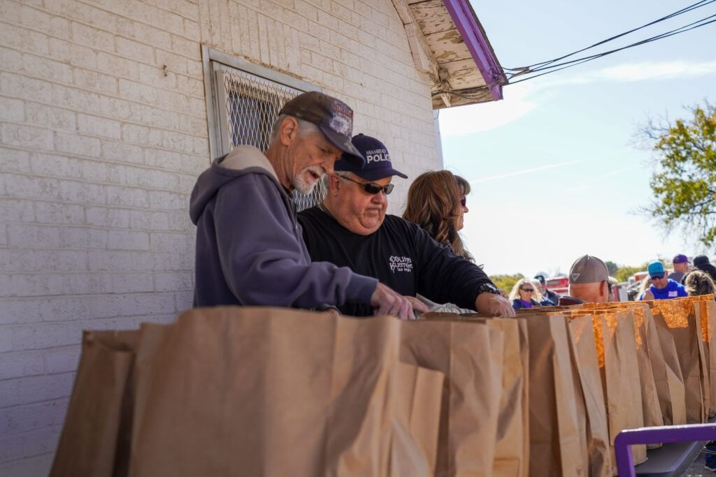 Three people leaning over a brown paper-covered table outdoors by a building, looking at something together.