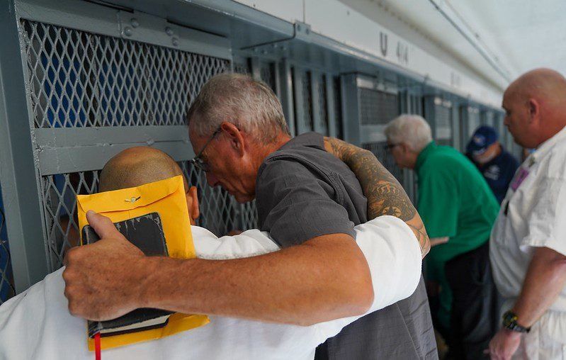 People hugging in a hallway, one holding a yellow tablet, with others waiting nearby.