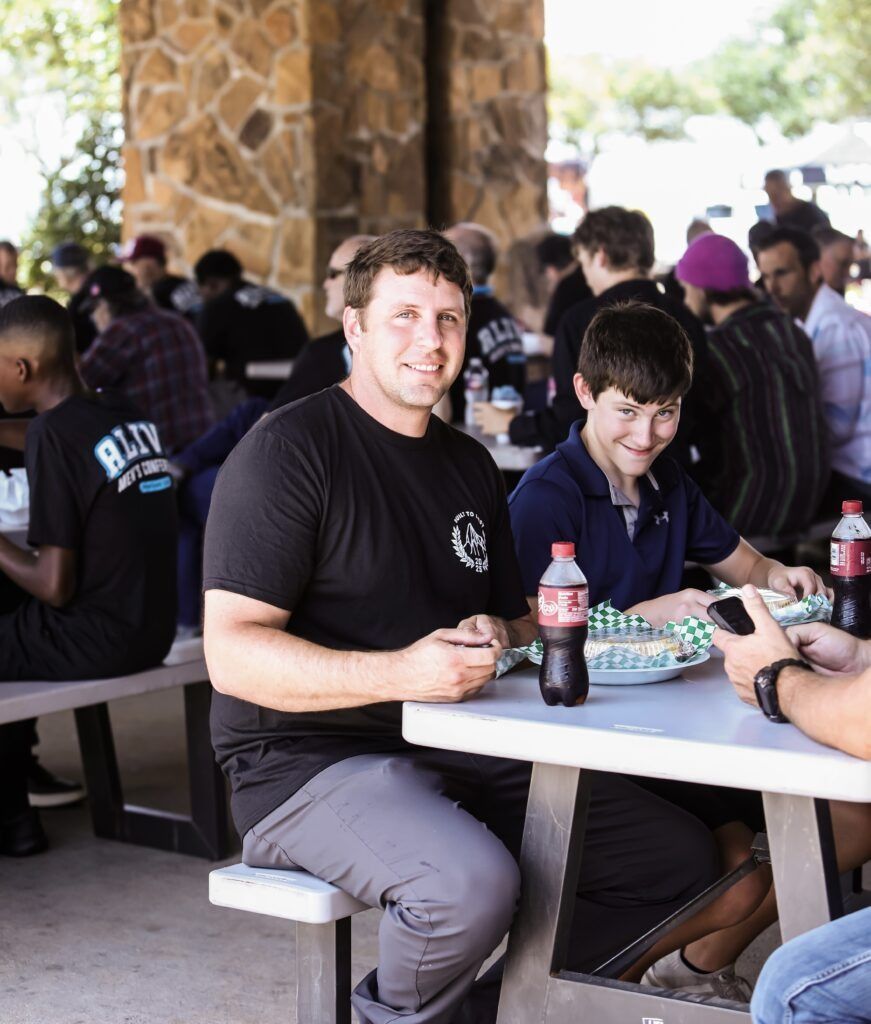 People seated at a picnic table outdoors, with drinks and food, in a crowded covered gathering area.