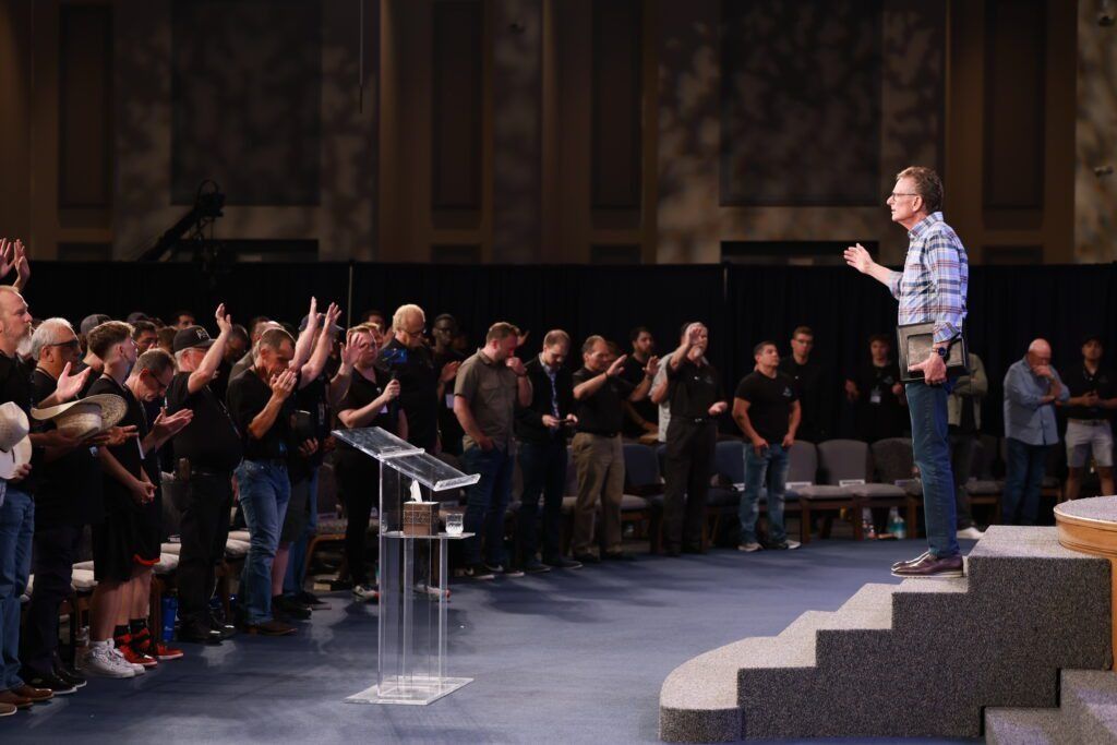 Speaker on stage addressing standing audience in a large auditorium, with hands raised in response.