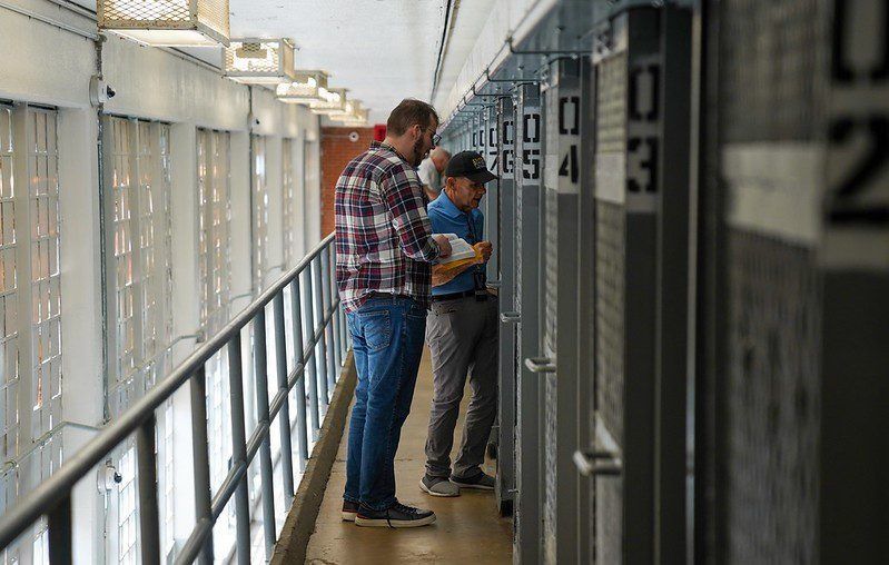 Two people standing in a narrow corridor, looking at papers near numbered doors or lockers