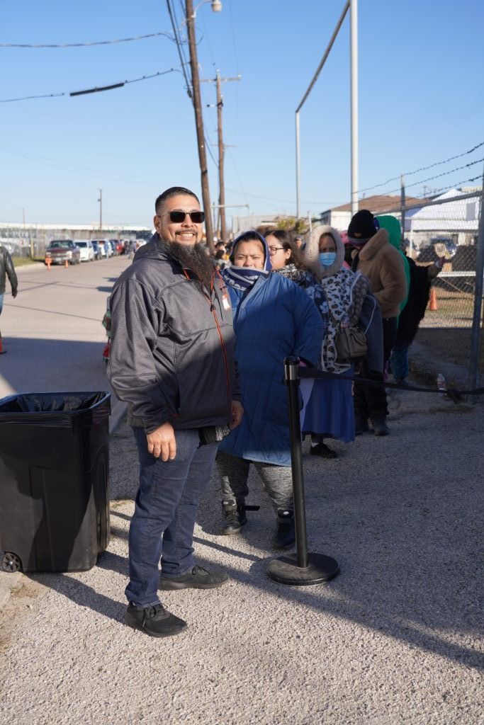 People standing in line outdoors beside a fence on a sunny day