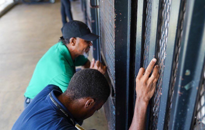 Two men in green and blue shirts peer through a barred window in a corridor