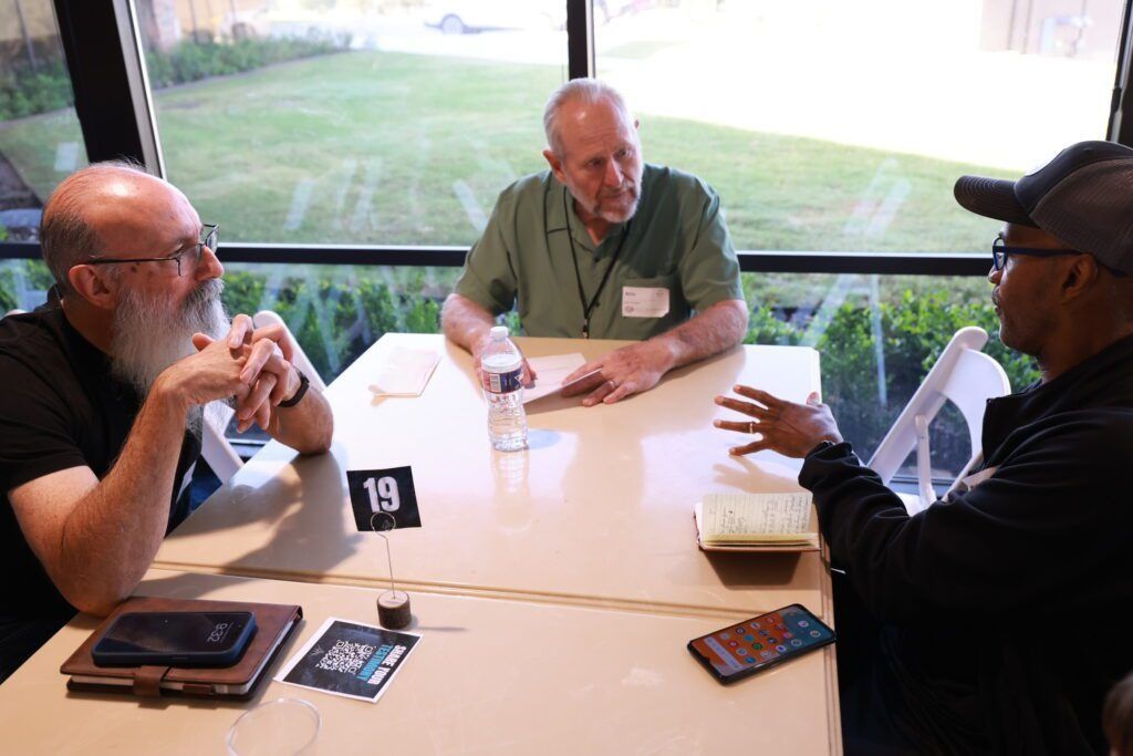 Three men talking at a round table with a number 19 sign, notebooks, and water bottles by a window